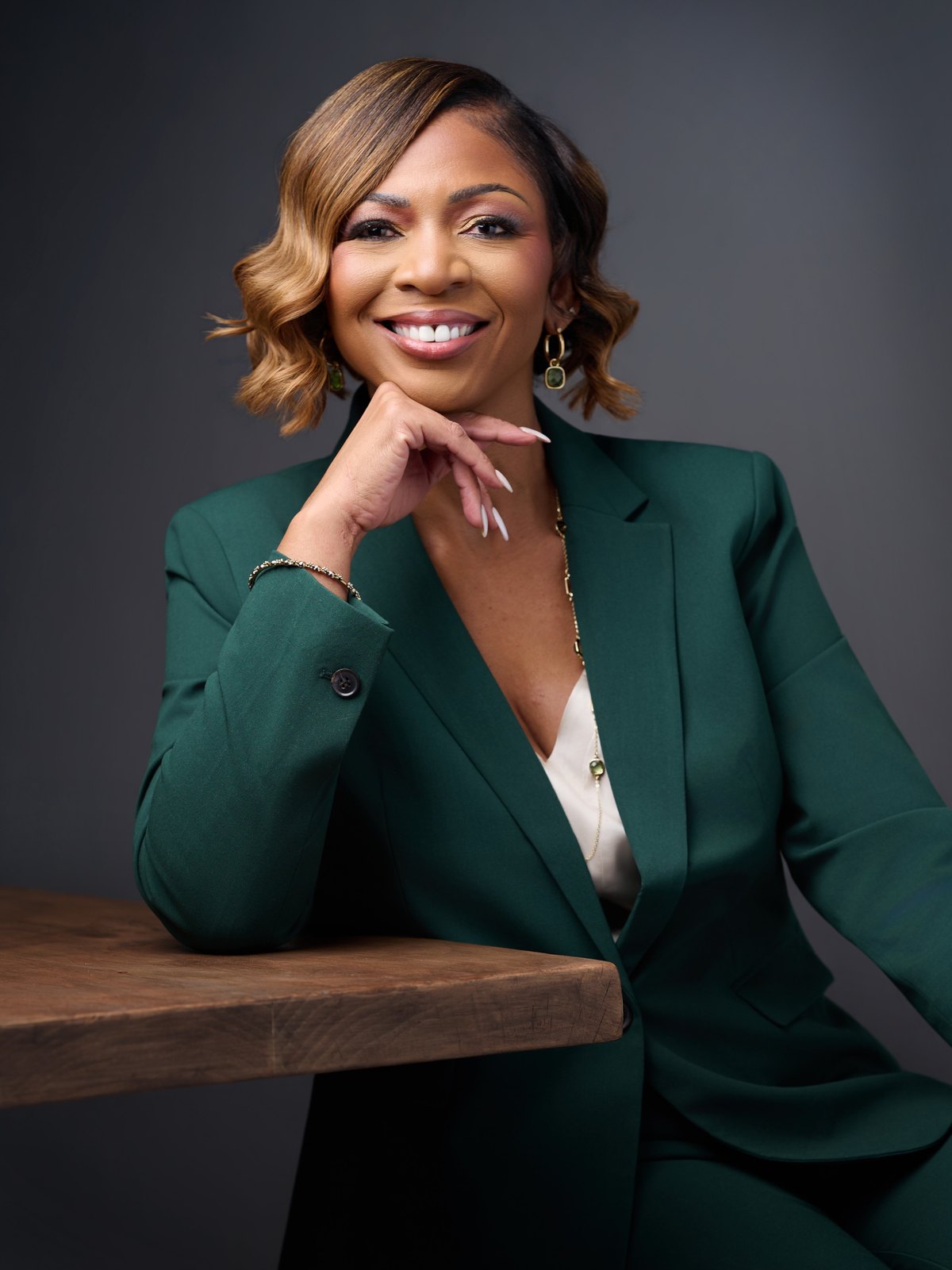 Heather Lynn, founder of Sovereign Advisory Co., in a deep green suit resting her chin on her hand at a wooden table against a soft gray studio backdrop
